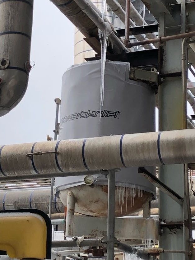 A large storage tank wrapped in a Powerblanket tank heating blanket, with icicles hanging from an overhead pipe rack