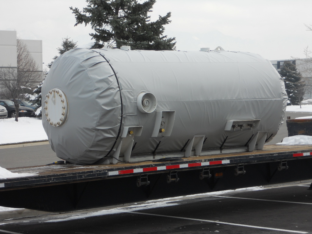 A large tank wrapped in an insulated Powerblanket cover, loaded on a flatbed truck in the snow for delivery