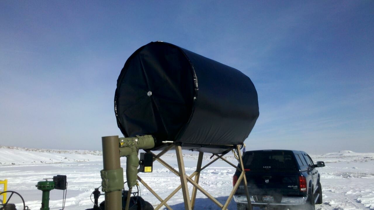 A 500-gallon tank wrapped in a Powerblanket heating blanket, mounted on a raised skid at a snow-covered well site